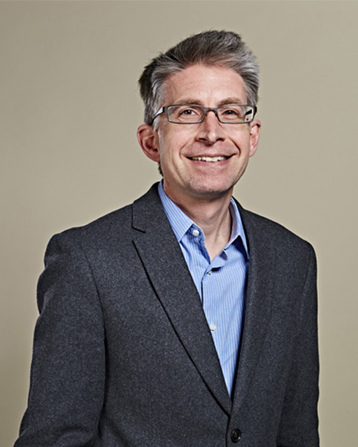 A man wearing glasses, a grey blazer and a blue shirt, smiling at the background
